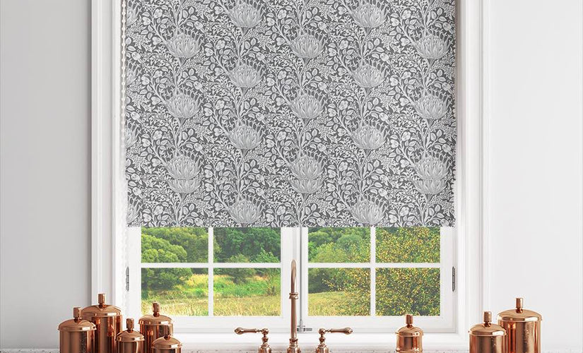 Charcoal patterned Roman blind fabric hanging in a kitchen window above blue cabinets with copper accessories