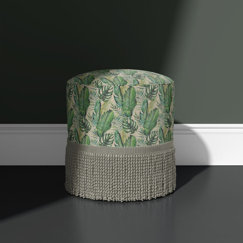 Amazon Leaf green upholstery fabric upholstered onto a round stool with fringe, shown against a dark green wall