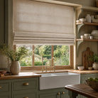 Broadway Plain Linen Fabric in natural beige shown as a tailored Roman blind in a traditional country kitchen with sage green cabinetry, oak worktops, farmhouse sink and soft natural daylight.
